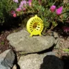 Piège à frelon OkyBee jaune posé sur des rochers avec des fleurs roses en arrière-plan